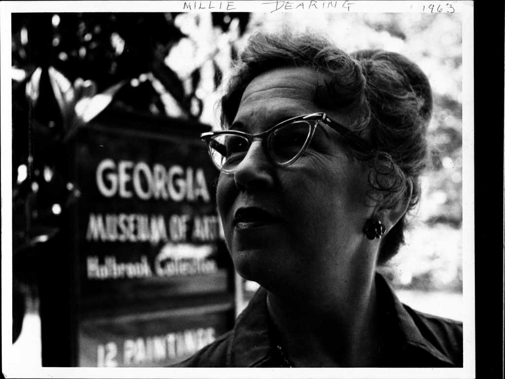 A portrait in grey scale of Millie Dearing with her hair in a bun and cat eye glasses standing beside the Georgia Museum of Art sign in 1963.
