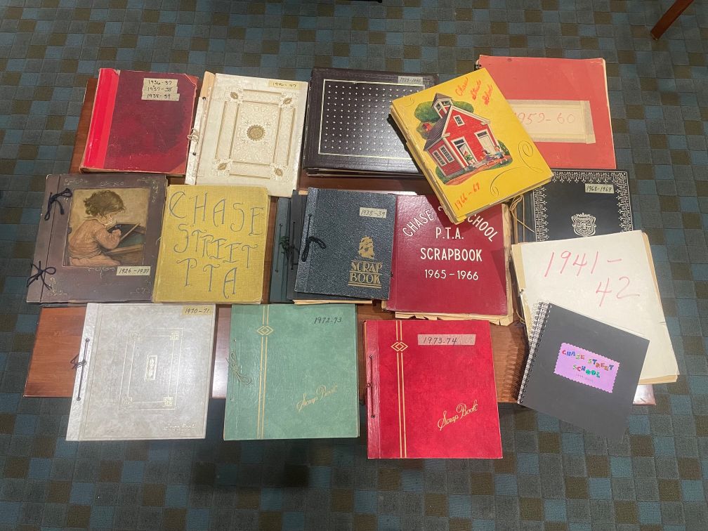 An overhead shot of 18 scrapbooks of varying sizes, colors, and years covering a table top. They are staggered and there is no apparent organization to how they are laid on the table. 